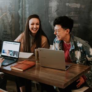 Three people are laughing around a table. On the table there are two laptops and a notebook.