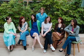 Zeven vrouwen zitten op een bankje in een groene omgeving, gekleed in verschillende stijlen en met blije gezichten.