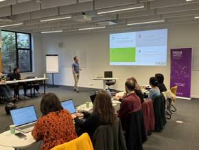 A presentation in a conference room featuring a speaker and an audience looking at laptops; slides display information and visuals.