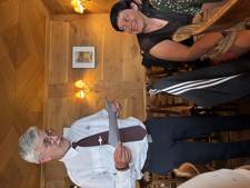 A waiter stands in a restaurant holding a menu while guests are seated at a table.