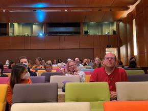 Two rows of people are sitting in a hall with colorful benches, some are wearing face masks.