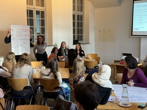 A group of people in a meeting room, with some participants giving a presentation while others listen and take notes.