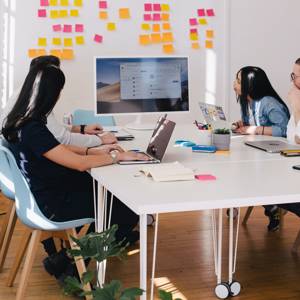 Five people in a meeting around a table all looking toward the screen that is on the table. Behind that screen are post-its on the wall.