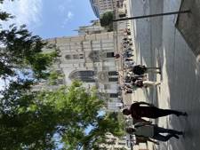 People are walking towards a large cathedral with a detailed façade and tall towers, surrounded by trees and a clear blue sky.