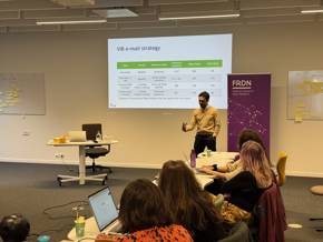 Presentation in a meeting room with a speaker in front of a screen, while attendees listen attentively.