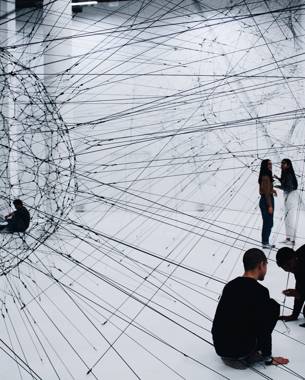 Photo of an artwork consisting of two translucent black spheres made from ropes. They are connected to each other like a kind of network. Further in the photo you can see that there are still people working on it.