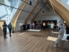 A group of people in a bright, modern room with wooden elements, engaged in interaction and discussion. Tables are arranged throughout the space.