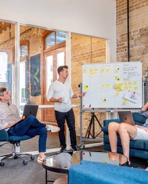 A presenter stands in front of a whiteboard on which a table has been drawn and post-its have been added into some cells. In the picture, three others are listening intently.