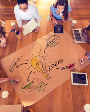 A group of people is working on a paper table cloth. On it a lightbulb is drawn and next to it it says the word 'Ideas' out of that arrows are drawn to the words